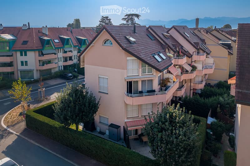 A multi-story residential building with balconies, surrounded by greenery and trees, with a clear view of the mountains in the background.