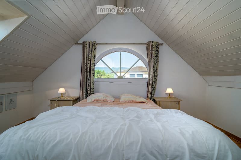 Cozy attic bedroom with slanted wooden ceilings, large arched window, bed with white duvet and colorful pillows, two nightstands with lamps, and patterned curtains.