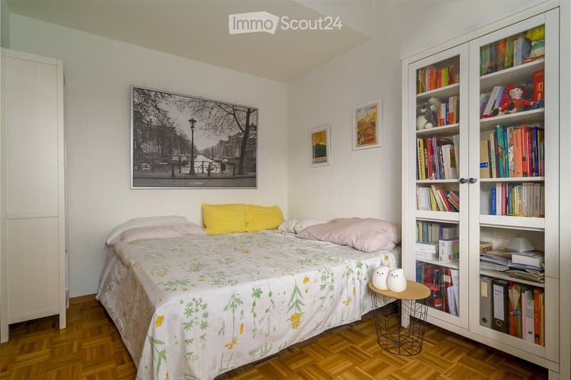Cozy bedroom with a single bed, colorful pillows, a large bookshelf filled with books and decorative items, a small round stool with two white ceramic figures, and framed artwork on the walls.