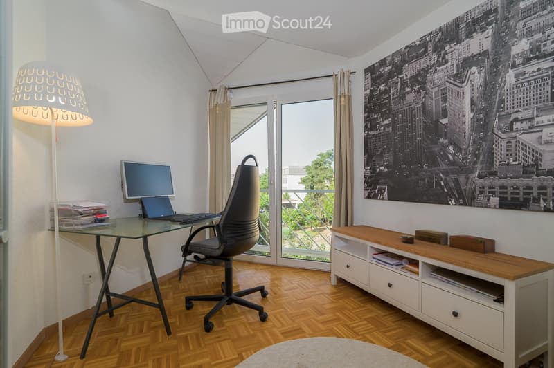 Modern office space with a glass desk, office chair, laptop, monitor, floor lamp, wooden cabinet, and a large cityscape picture on the wall. The room features wooden flooring and large windows with a view of a balcony.