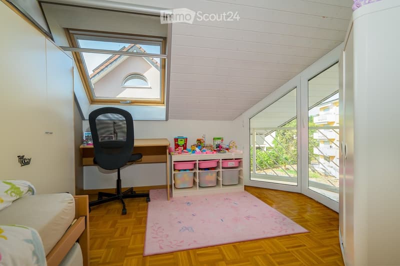 Bright bedroom with wooden flooring, slanted white ceiling, large window, and open balcony door leading to a terrace. Contains a bed, desk with chair, storage unit with bins, and a pink rug.