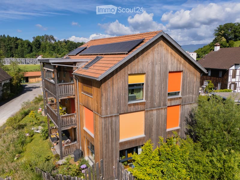 Modern two-story house with wooden exterior, solar panels on the roof, and a balcony with outdoor furniture.