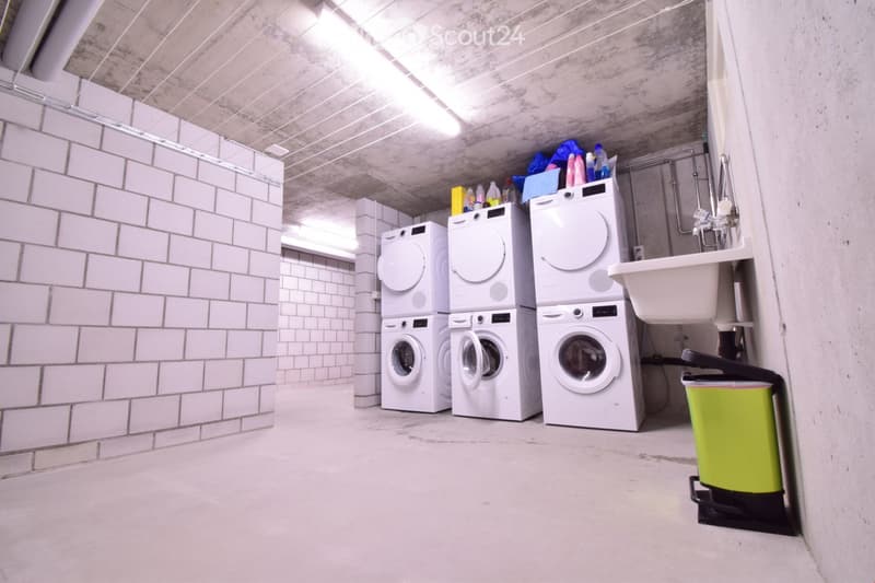 Clean, modern laundry area with white tiled walls, concrete ceiling, and three stacked washing machines. Includes a sink with a faucet and a green trash bin.