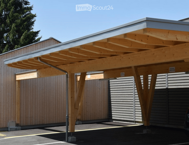 Carport with wooden frame and metal roof, attached to a building with wooden siding and grey paneling.