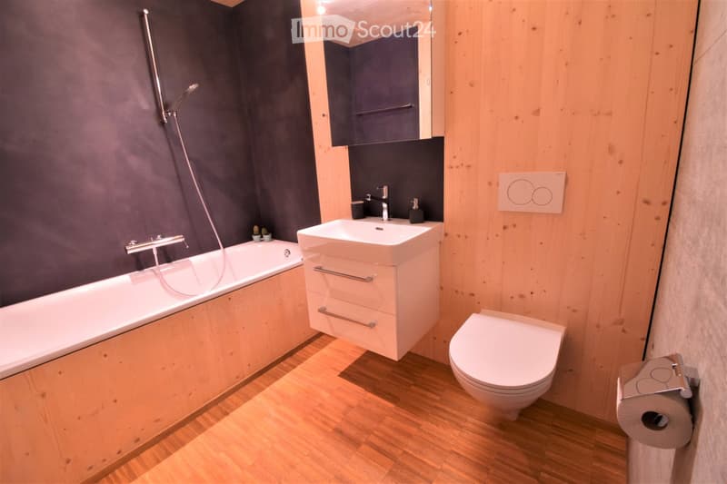 Modern bathroom with a bathtub, sink, toilet, and mirror. Features wooden walls and flooring, with a dark accent wall and a sleek, contemporary design.