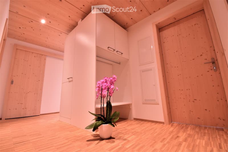 A small, modern hallway with wooden flooring, white walls, and a built-in wardrobe. A potted orchid is placed on the floor near the wardrobe.