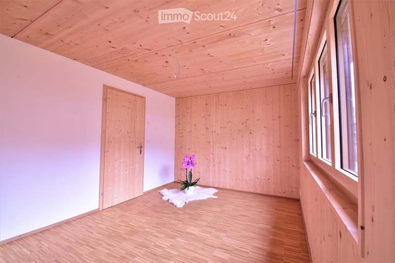 Empty room with wooden floors, light pink walls, and a wooden ceiling. Features include a door, windows, and a small white rug with a flower vase.