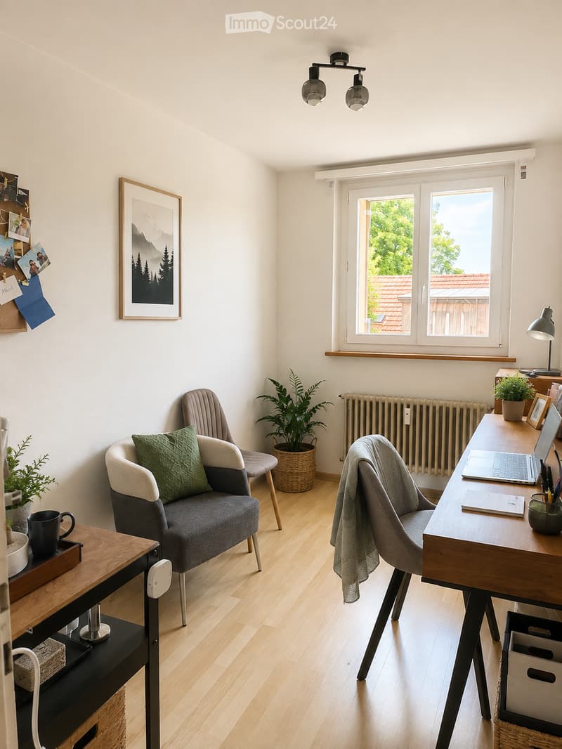 A modern, well-lit room with a sofa, armchair, desk, laptop, and decorative plants. Features large windows with a view of greenery outside, a radiator, and a picture frame on the wall.