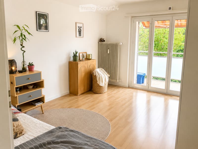 Bright bedroom with wooden flooring, large sliding glass doors leading to a balcony, a bed with grey bedding, a nightstand with a lamp, a drawer unit, a wooden cabinet, a wicker basket, and decorative plants.