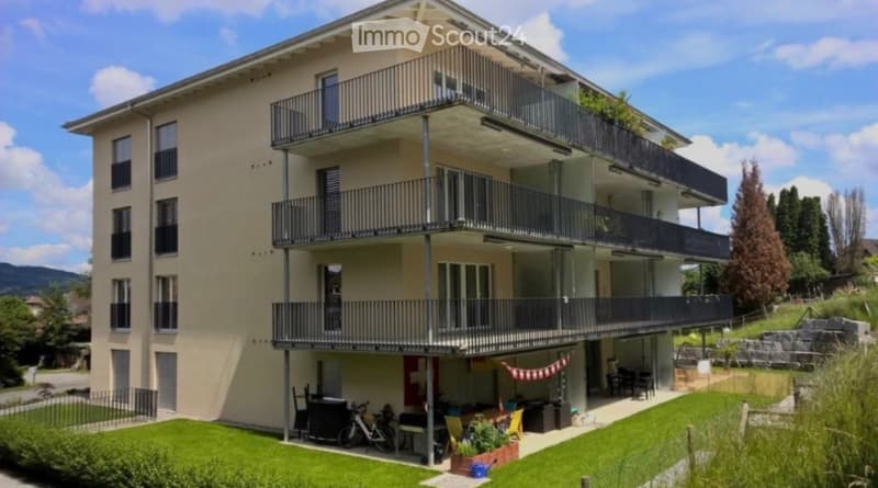Modern three-story apartment building with multiple balconies, surrounded by greenery and a small garden area with outdoor furniture.