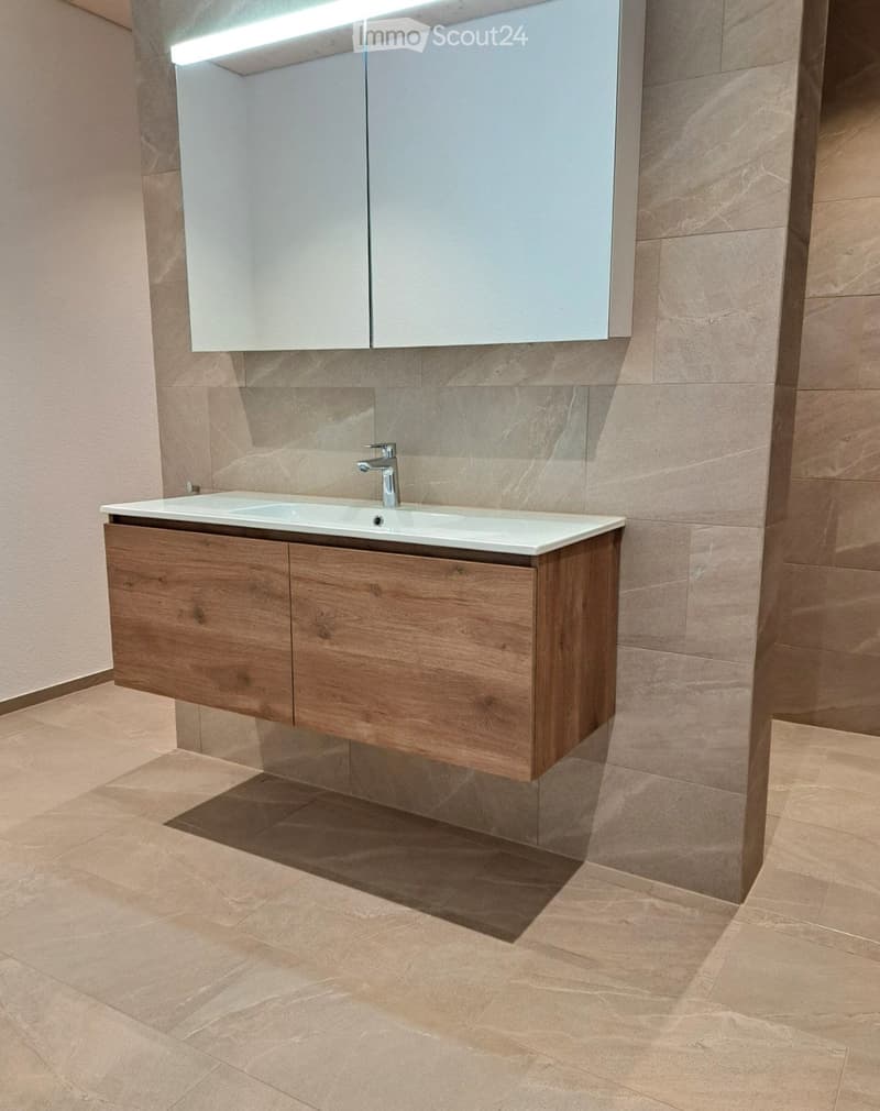 Modern bathroom with a double sink, wooden cabinet, and large mirror above it. The walls and floor are tiled in a light beige color.