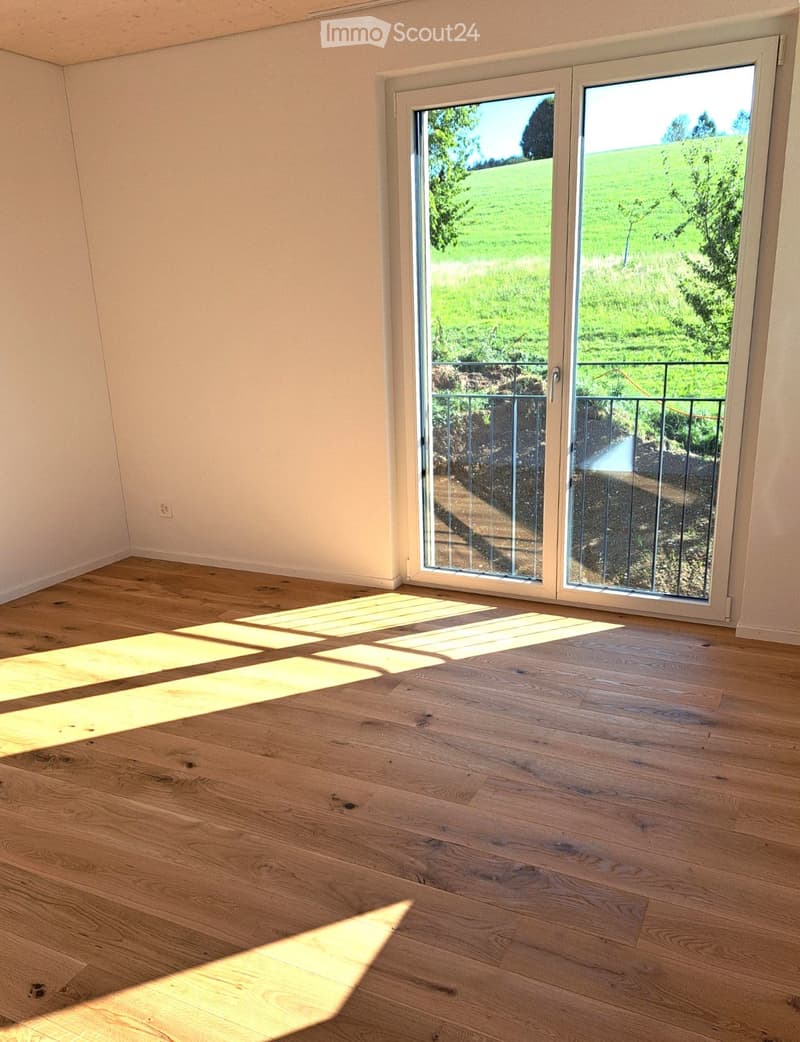 Empty room with wooden flooring and large sliding glass doors leading to a balcony with a view of a green field.