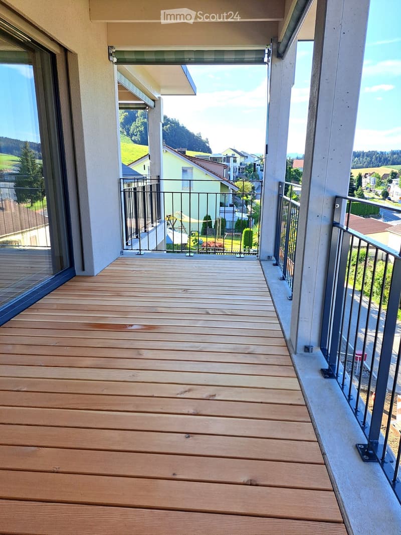 Wooden deck balcony with metal railing, overlooking a residential area with houses and greenery.