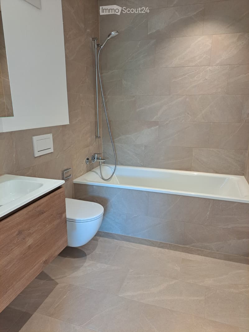Modern bathroom with a bathtub, toilet, sink, and showerhead. The walls are tiled in a marble-like pattern, and the floor is tiled in a light beige color.