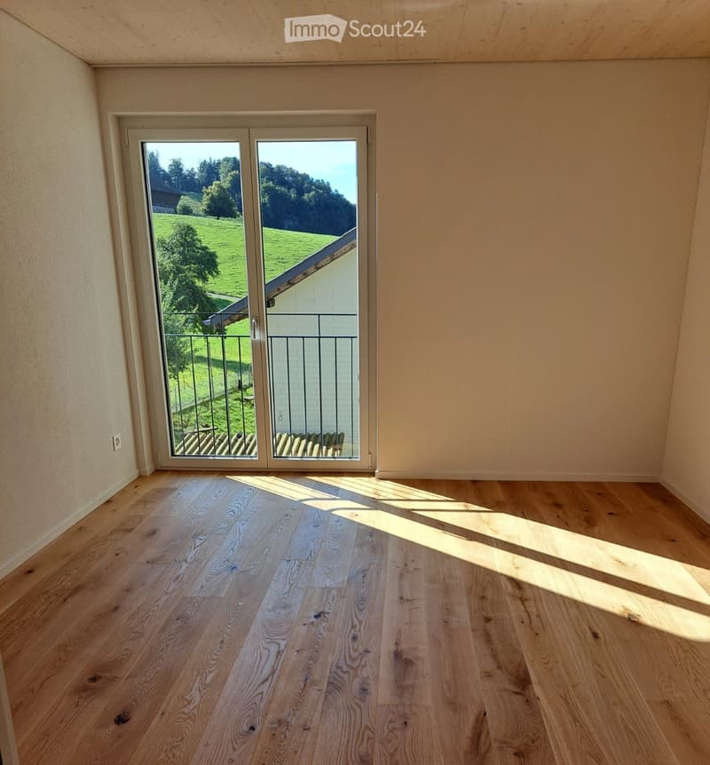 Empty room with wooden flooring and large sliding glass doors leading to a balcony with a scenic view of greenery and a house.