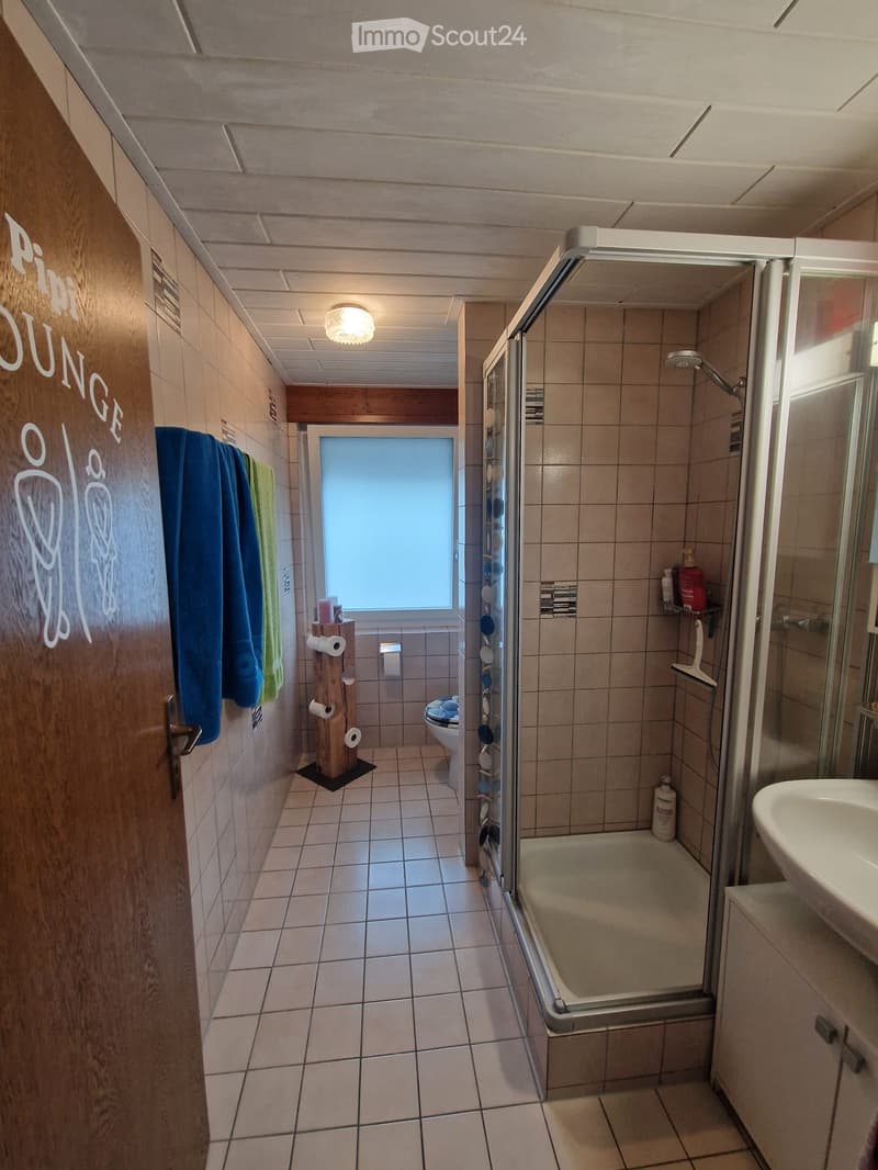 Modern bathroom with tiled walls and floor, featuring a shower stall, toilet, sink, and towel rack. The space includes a window for natural light and a decorative wooden toilet paper holder.