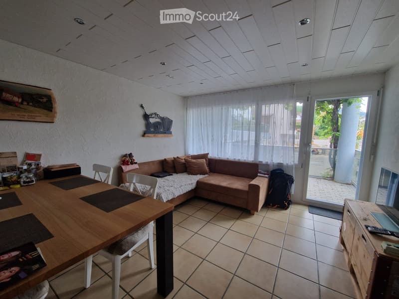 A cozy living room with a beige sofa, wooden dining table, white chairs, and a backpack near the sliding glass door. The room features tiled flooring, white walls, and a large window with sheer curtains.