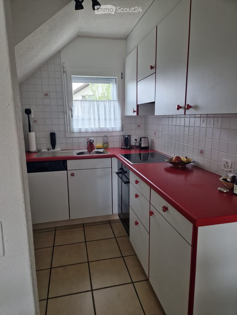 Compact kitchen with red countertops, white cabinets, tiled walls, and a window with a curtain. Includes a sink, stove, oven, and various small appliances.