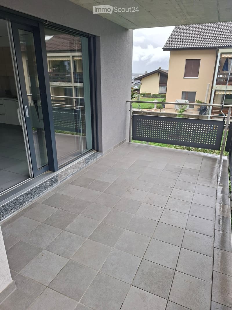 Modern balcony with large grey tiles, glass sliding door, metal railing, and view of neighboring houses.