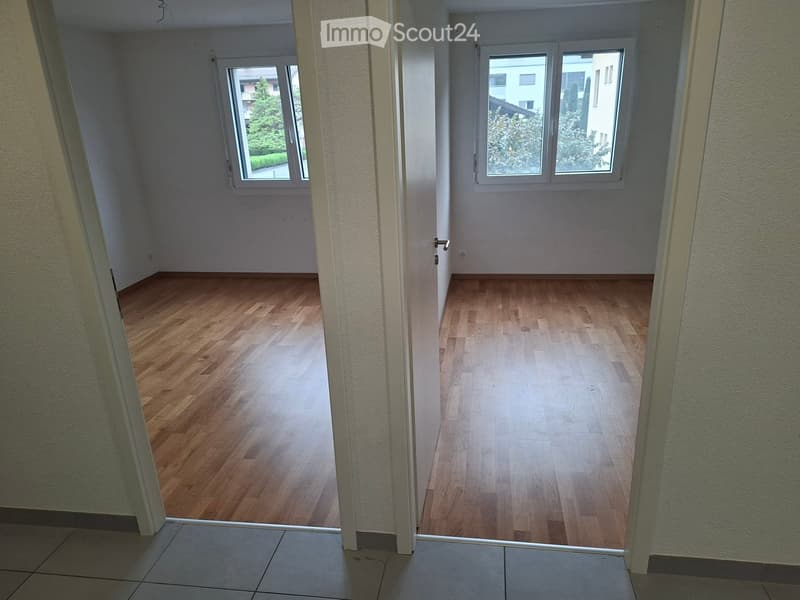 Empty room with wooden flooring, white walls, and large windows allowing natural light. The room is unfurnished and appears to be part of a larger space.