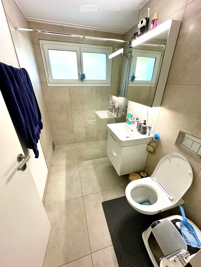 Modern bathroom with beige tiles, a white sink, toilet, mirror, and a shower stall. A dark blue towel hangs on the wall, and a small black mat is placed in front of the toilet.