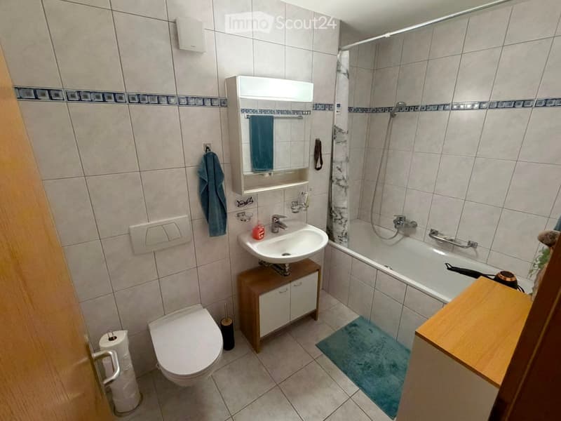 Modern bathroom with a toilet, sink, bathtub, mirror cabinet, and a towel hanging on the wall. The floor is tiled, and there is a small wooden cabinet under the sink.