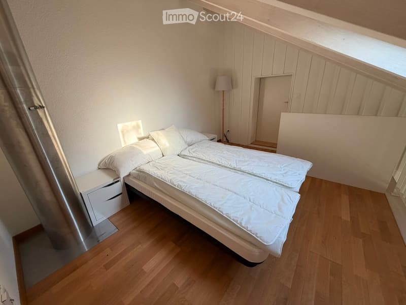 Cozy bedroom with a white bed, nightstand, and floor lamp. Wooden flooring and white walls.