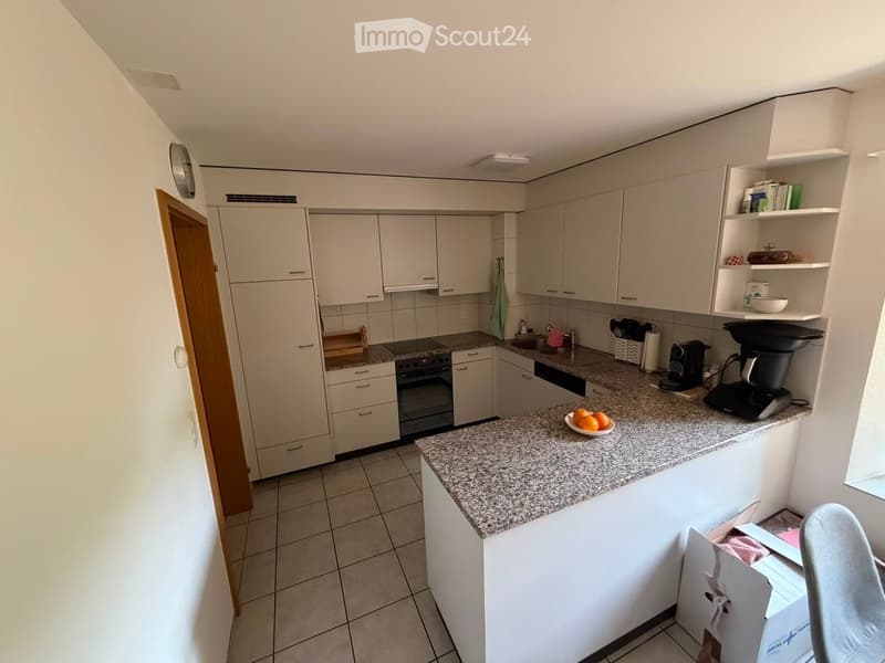 Modern kitchen with white cabinets, granite countertop, built-in oven, dishwasher, and coffee maker. Includes a bowl of oranges on the counter.