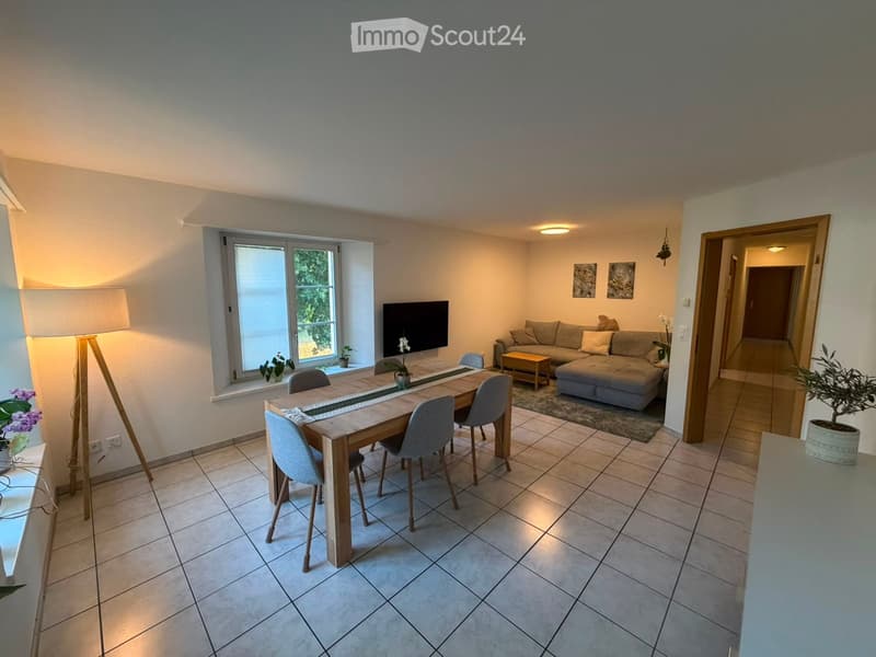 Bright living and dining area with tiled flooring, large window, sofa, coffee table, dining table with chairs, and television mounted on wall.
