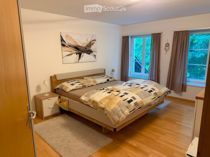 Modern bedroom with wooden flooring, large bed with patterned bedding, nightstand with lamp, large windows with curtains, and a decorative painting on the wall.
