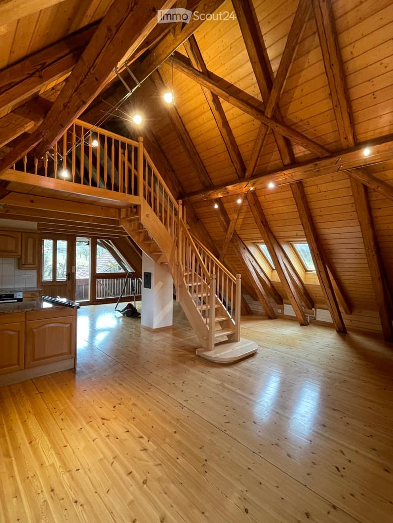 Spacious attic room with wooden beams, wooden floor, and a wooden staircase leading to an upper level. Large windows provide natural light.