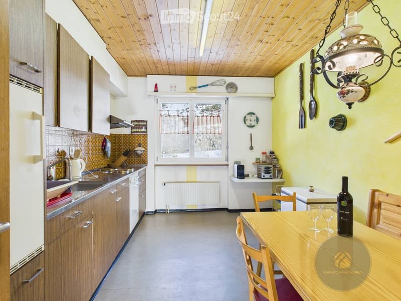 Bright kitchen with wooden cabinets, a central dining table with chairs, and various appliances including a refrigerator, sink, oven, and microwave. A chandelier hangs from the ceiling, and decorative items are displayed on the walls and shelves.