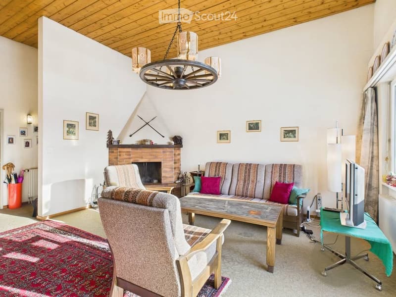 Cozy living room with a fireplace, wooden ceiling, and large windows. Features a sofa, armchair, coffee table, television, and decorative picture frames.