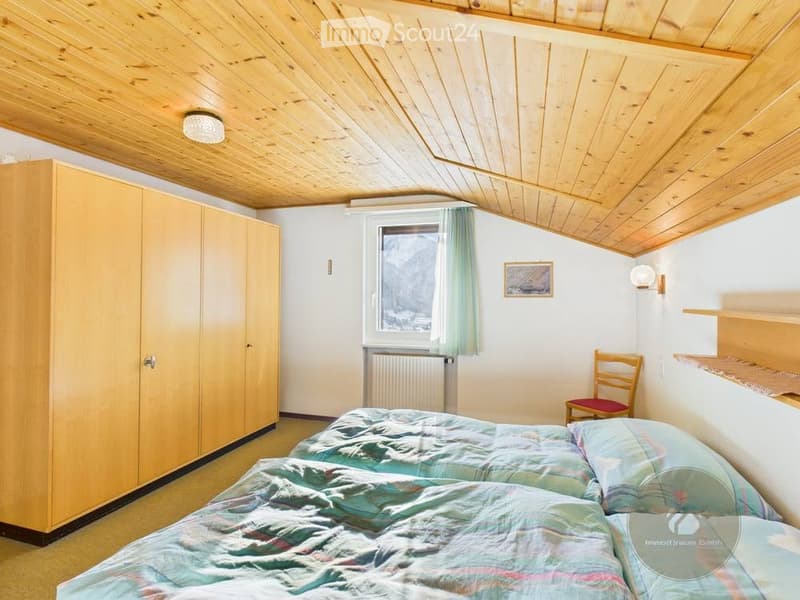 Cozy bedroom with wooden slanted ceiling, two single beds, wardrobe, small chair, radiator, and a view through the window.