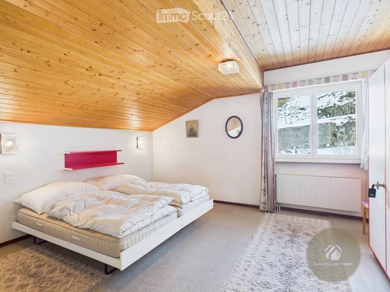 Cozy bedroom with a white bed, wooden slanted ceiling, large window with view, red shelf, and a small round mirror on the wall.