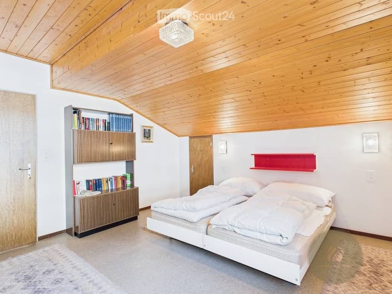 Cozy bedroom with wooden ceiling, white walls, a double bed, a wooden bookshelf filled with books, a red shelf, and a picture frame on the wall.