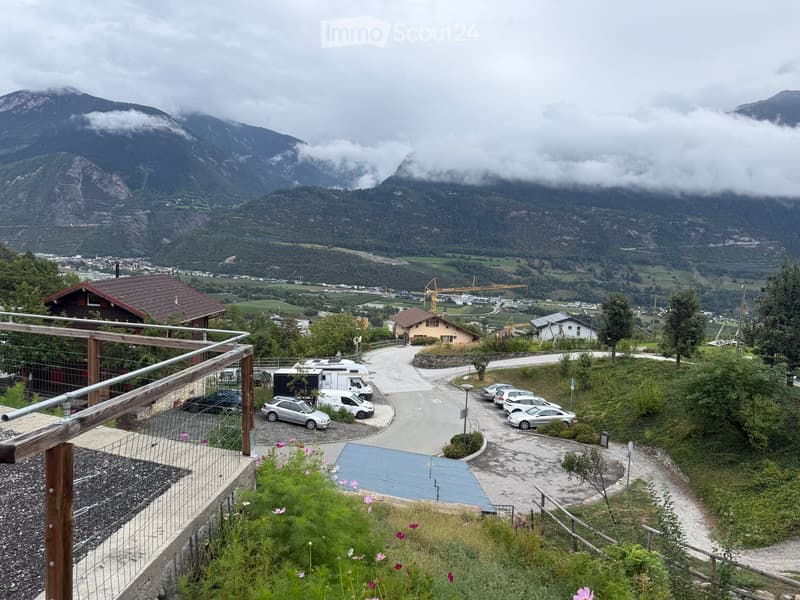 A scenic outdoor view from a balcony or terrace overlooking a valley with houses, parked cars, and distant mountains partially covered in clouds.