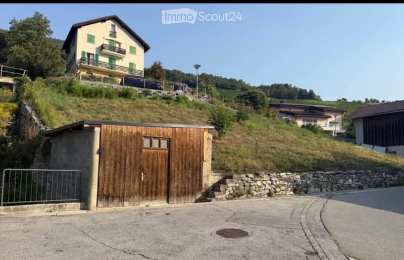 A two-story house with green shutters and balconies sits on a hillside, with a wooden garage at the base of the slope.