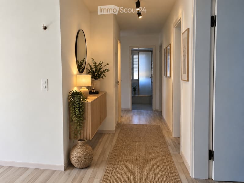 Modern hallway with wooden flooring, white walls, and soft lighting. Features a small wooden console table with a lamp, decorative plants, and a woven rug leading to an open doorway.