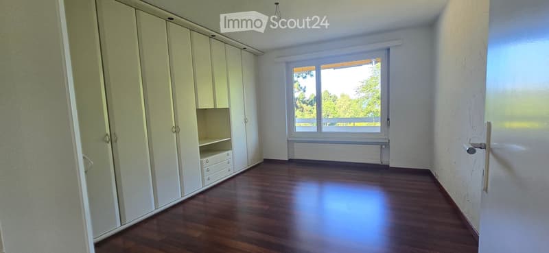 Empty room with wooden flooring, white walls, large window with view, built-in wardrobe with drawers, and radiator under the window.