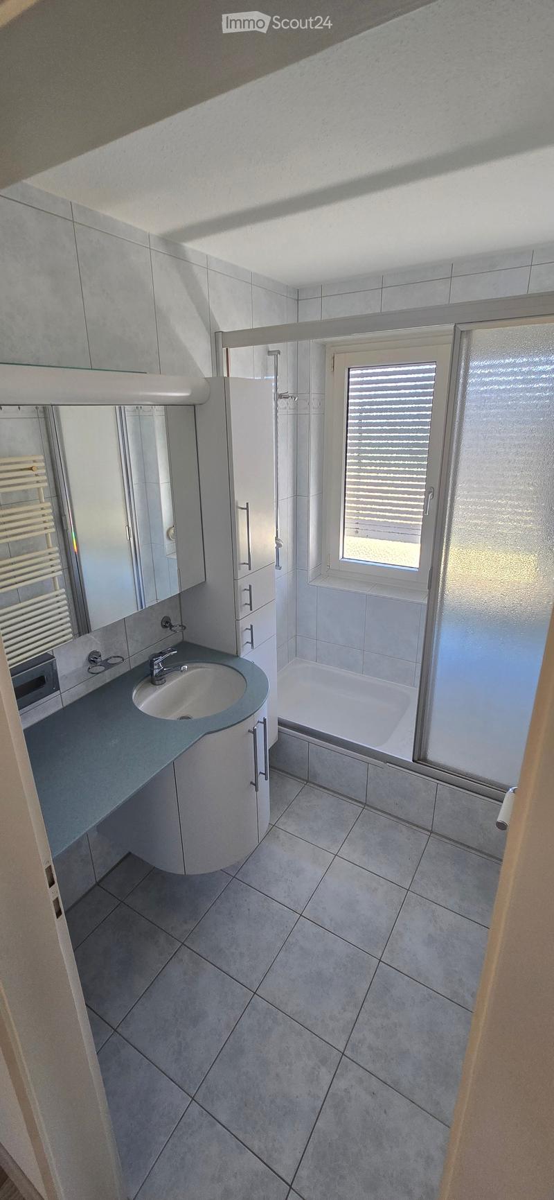 Modern bathroom with a sink, mirror, bathtub, and radiator. The space features tiled walls and floor, a window with blinds, and a sleek cabinet with a countertop.