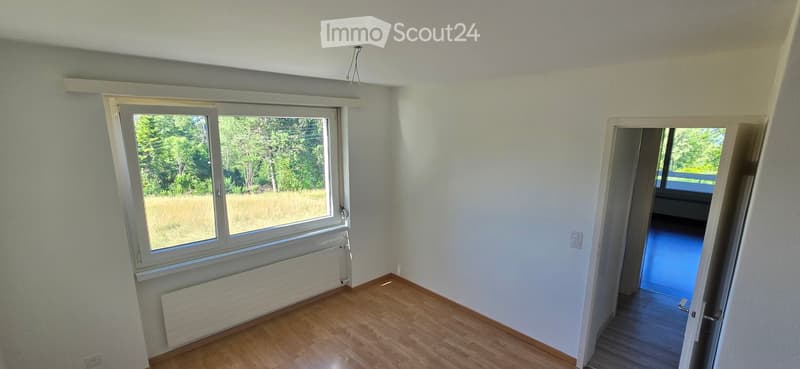 Empty room with wooden flooring, large window, radiator, and an open doorway leading to another room.