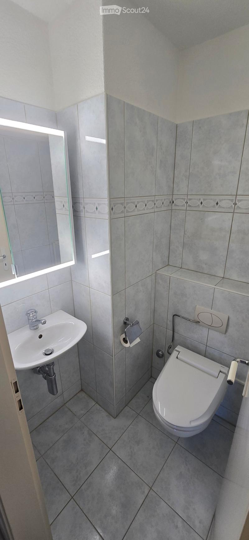 Modern bathroom with grey tiled walls and floor, featuring a white sink with a chrome faucet, a toilet with a toilet paper holder, and a mirror with built-in lighting.