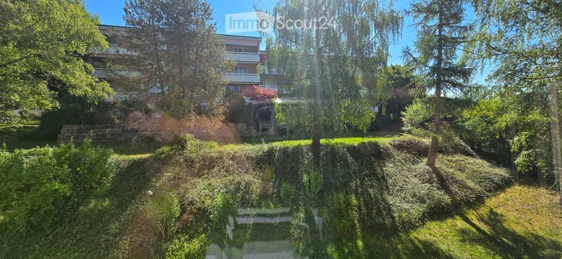 A well-maintained garden area with lush greenery, trees, and a multi-story building in the background.