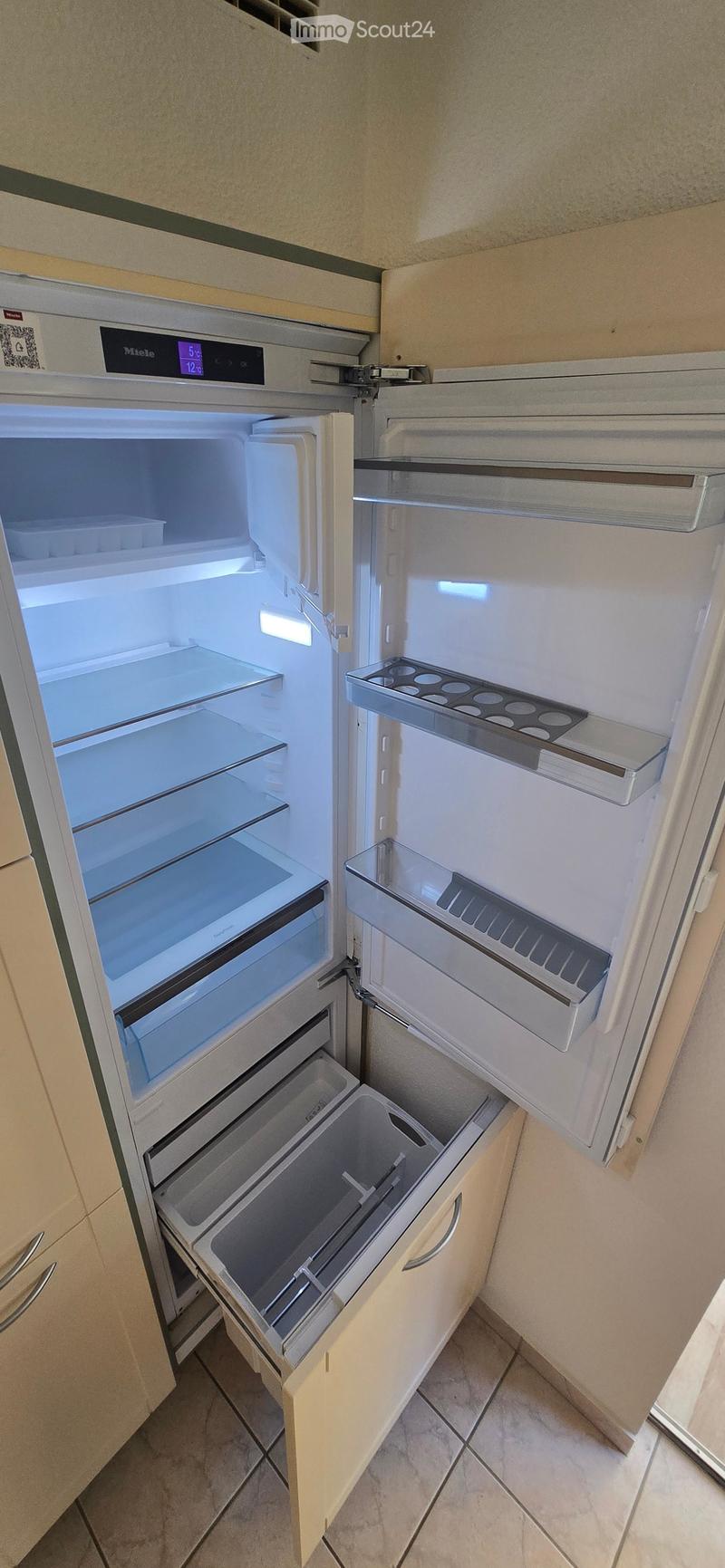 Open refrigerator with multiple compartments and shelves, integrated into a modern kitchen with light-colored cabinets and tiled flooring.