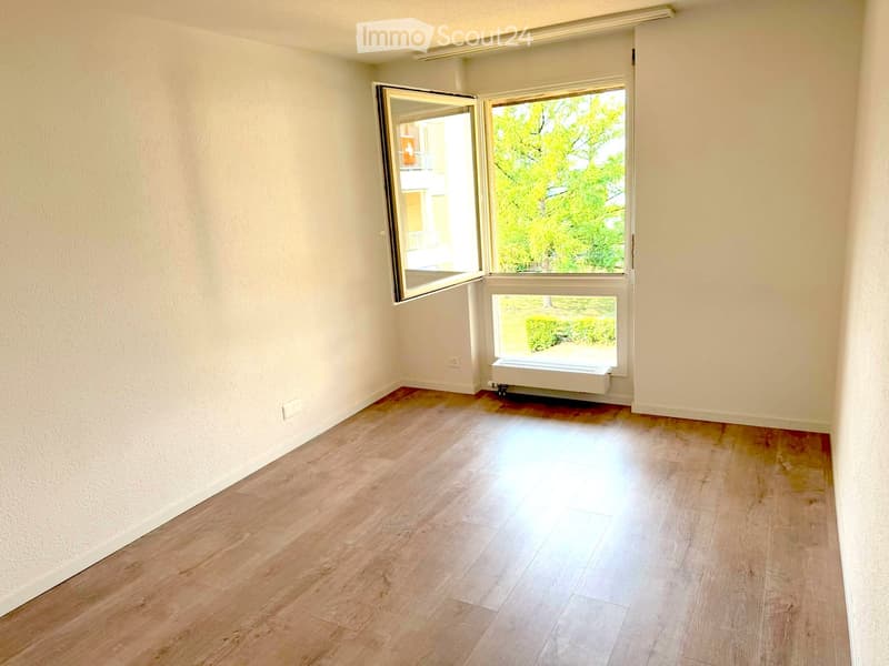 Empty room with light wooden flooring, white walls, and large windows with blinds, offering a view of greenery outside.