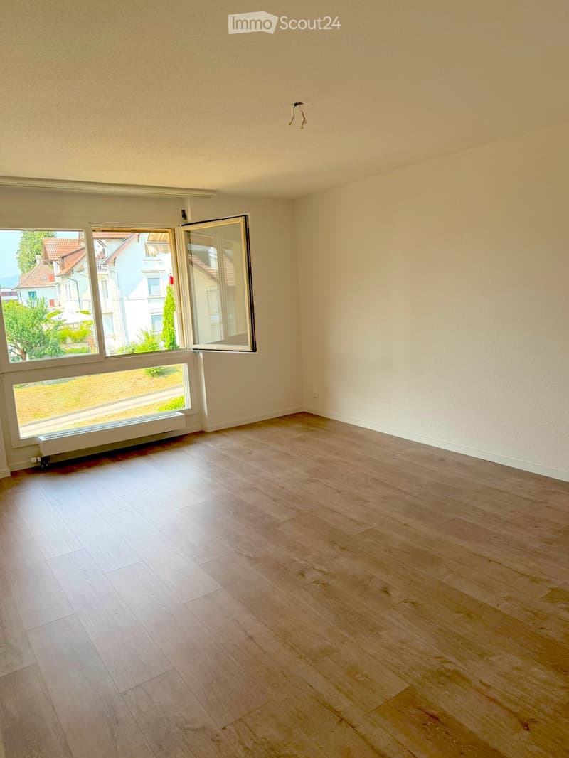 Empty room with light wooden flooring, white walls, and large windows offering a view of the street and neighboring houses.