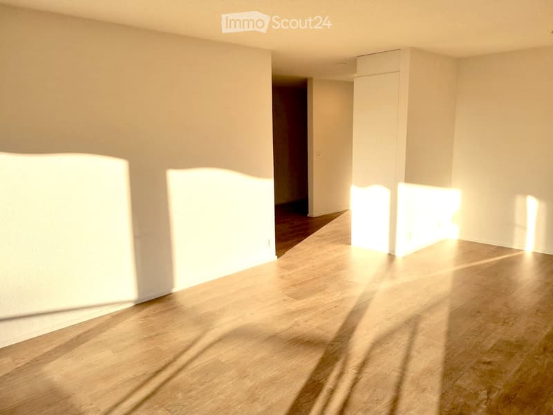 Empty room with light wooden flooring and white walls, featuring natural light from a window and an open doorway leading to another room.