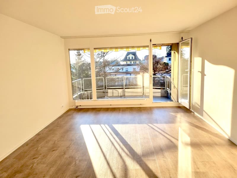 Empty room with light wooden flooring, large windows, and an open sliding door leading to a balcony area.