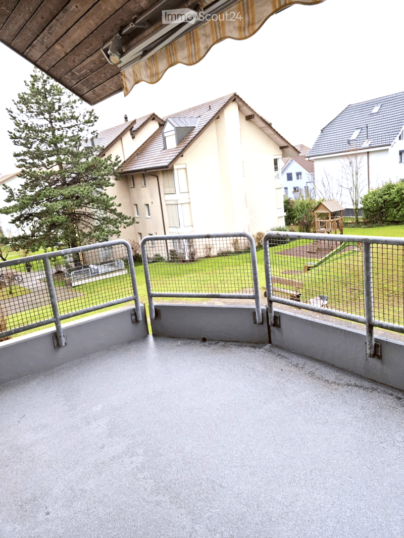 Empty balcony with metal railing, concrete floor, and a view of a residential area with houses, trees, and a playground.